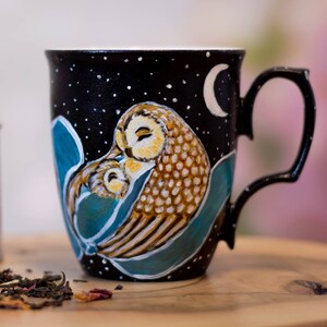 Könnte beinhalten: Eine schwarze Keramiktasse mit handbemaltem Design von zwei Eulen, die sich auf einem Ast gegen einen sternenklaren Nachthimmel aneinanderkuscheln. Die Tasse hat einen weißen Halbmond und weiße Sterne, die auf den schwarzen Hintergrund gemalt sind. Die Eulen sind in Braun-, Gelb- und Weißtönen bemalt. Die Tasse hat einen weißen Henkel.