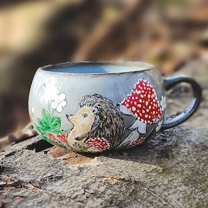 May include: A gray ceramic mug with a hand-painted design of a hedgehog and a red mushroom. The mug is sitting on a tree trunk.