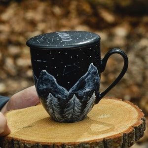 Può includere: Una tazza in ceramica nera con una scena di montagna bianca e stelle dipinte su di essa. La tazza ha un coperchio e un manico. La tazza è appoggiata su una fetta di legno.