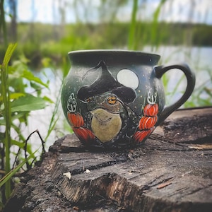 May include: A dark green ceramic mug with a whimsical frog design wearing a witch's hat. The mug features a handle and is decorated with orange pumpkins and a full moon. The mug is set on a weathered tree stump, with a natural background.
