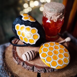 May include: A black mug with a honeycomb pattern, a jar of honey with a burlap lid, a wooden honey dipper, and a yellow and white honeycomb patterned lid on a wooden slice.