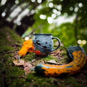 May include: A Halloween-themed mug with a witch and spiderweb design, alongside two decorative gourds in orange and green hues. The mug is dark blue with a handle. The scene is set on a mossy surface, evoking an autumnal atmosphere.