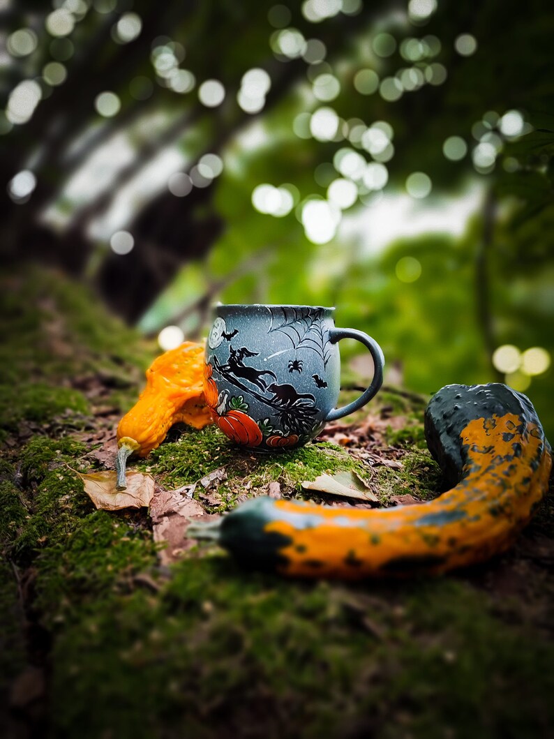 May include: A Halloween-themed mug with a witch and spiderweb design, alongside two decorative gourds in orange and green hues. The mug is dark blue with a handle. The scene is set on a mossy surface, evoking an autumnal atmosphere.