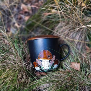 May include: A black ceramic mug with a brown rim and a hand-painted design of mushrooms. The mug is sitting in a bed of green grass and brown leaves.