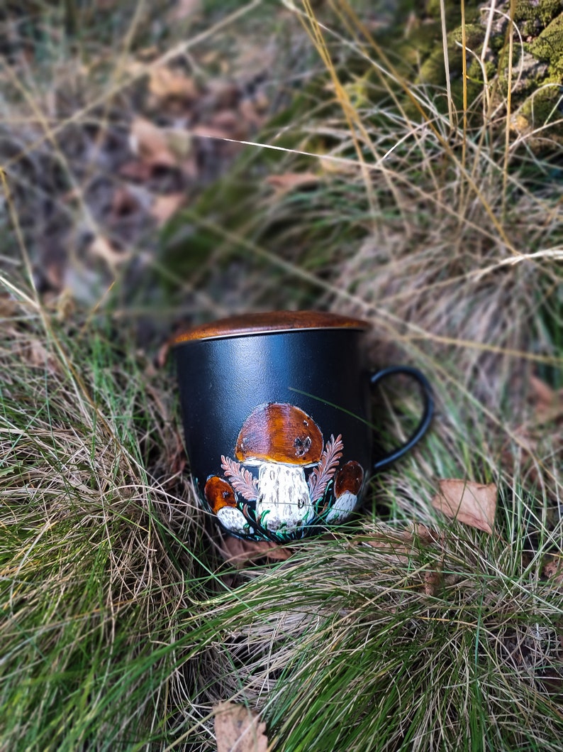 May include: A black ceramic mug with a brown rim and a hand-painted design of mushrooms. The mug is sitting in a bed of green grass and brown leaves.