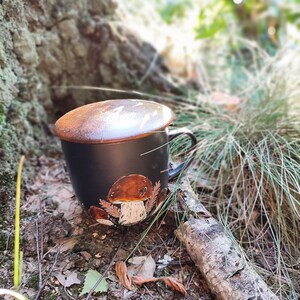 May include: Black ceramic mug with a mushroom design. The mug has a brown lid and is sitting on a bed of leaves and twigs.