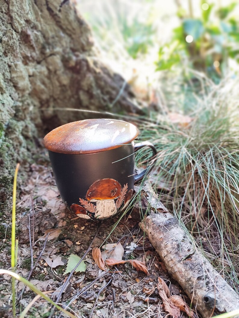 May include: Black ceramic mug with a mushroom design. The mug has a brown lid and is sitting on a bed of leaves and twigs.