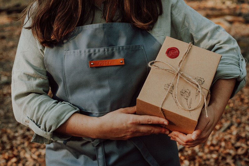 May include: A person wearing a grey apron with a leather name tag that says "Justine" is holding a brown gift box wrapped with twine and a red wax seal. The box has a floral design.