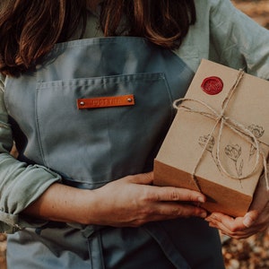 May include: A gray apron with a leather tag that reads "JUSTYNA" is held by a person. The person is also holding a brown cardboard box tied with twine and sealed with a red wax stamp. The box has a floral design.