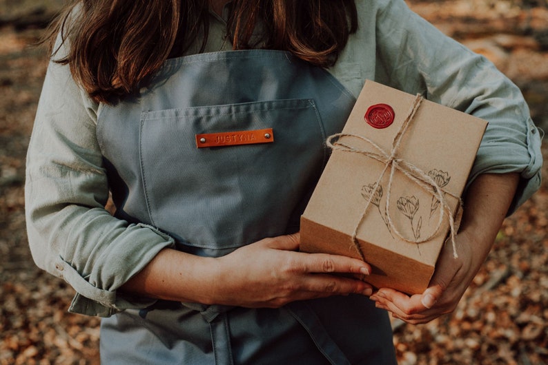 May include: A gray apron with a leather tag that reads "JUSTYNA" is held by a person. The person is also holding a brown cardboard box tied with twine and sealed with a red wax stamp. The box has a floral design.