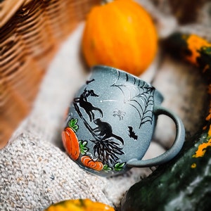 May include: A gray ceramic Halloween mug with a black cat riding a broom, pumpkins, and a spiderweb design. The mug is surrounded by pumpkins and gourds, with a knitted blanket in the background.