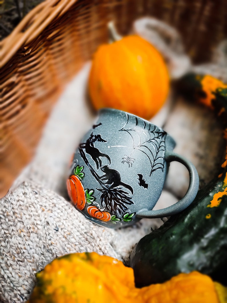 May include: A gray ceramic Halloween mug with a black cat riding a broom, pumpkins, and a spiderweb design. The mug is surrounded by pumpkins and gourds, with a knitted blanket in the background.