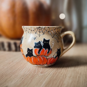 May include: A ceramic mug with a brown and beige speckled glaze. The mug features a hand-painted design of black cats sitting on orange pumpkins with a spider web in the background.