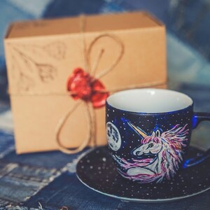 Peut inclure: Un ensemble tasse et soucoupe en c&eacute;ramique peint &agrave; la main avec un motif de licorne sur un fond bleu fonc&eacute; &eacute;toil&eacute;. La tasse a un int&eacute;rieur blanc et une soucoupe assortie. Une bo&icirc;te cadeau marron avec un sceau de cire rouge est en arri&egrave;re-plan.