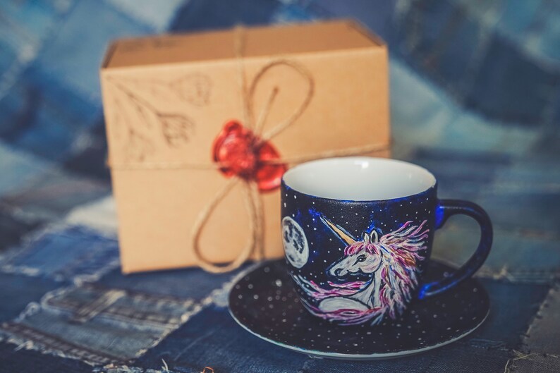 Peut inclure: Un ensemble tasse et soucoupe en c&eacute;ramique peint &agrave; la main avec un motif de licorne sur un fond bleu fonc&eacute; &eacute;toil&eacute;. La tasse a un int&eacute;rieur blanc et une soucoupe assortie. Une bo&icirc;te cadeau marron avec un sceau de cire rouge est en arri&egrave;re-plan.