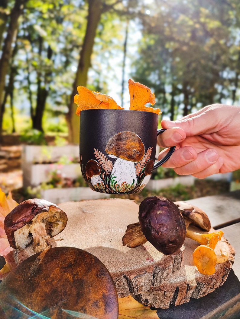 May include: A black ceramic mug with a mushroom design and a hand holding the mug. The mug is filled with orange chanterelle mushrooms. The mug is sitting on a wooden surface with other mushrooms.