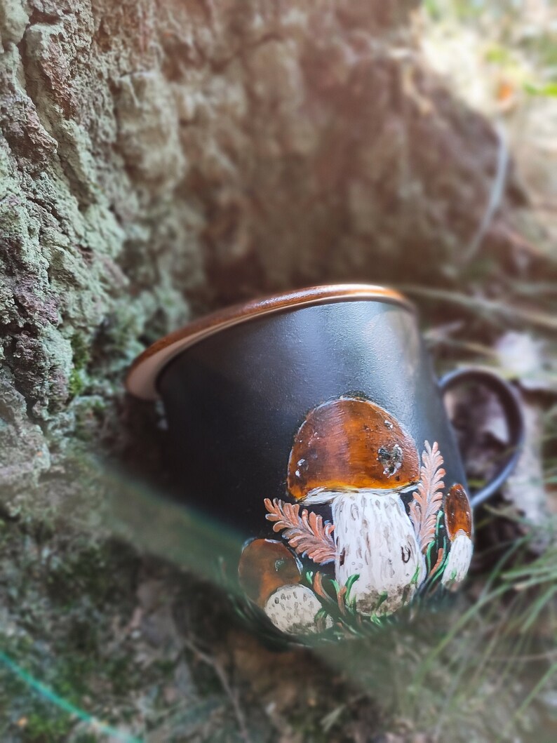 May include: Black ceramic mug with a brown rim and a hand-painted design of mushrooms. The mushrooms are brown, white, and orange with green leaves.