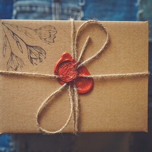 Peut inclure: Une bo&icirc;te cadeau rectangulaire marron attach&eacute;e avec de la ficelle et scell&eacute;e avec un sceau de cire rouge. La bo&icirc;te a un motif floral dans le coin sup&eacute;rieur gauche. Le fond est un tissu en denim bleu.