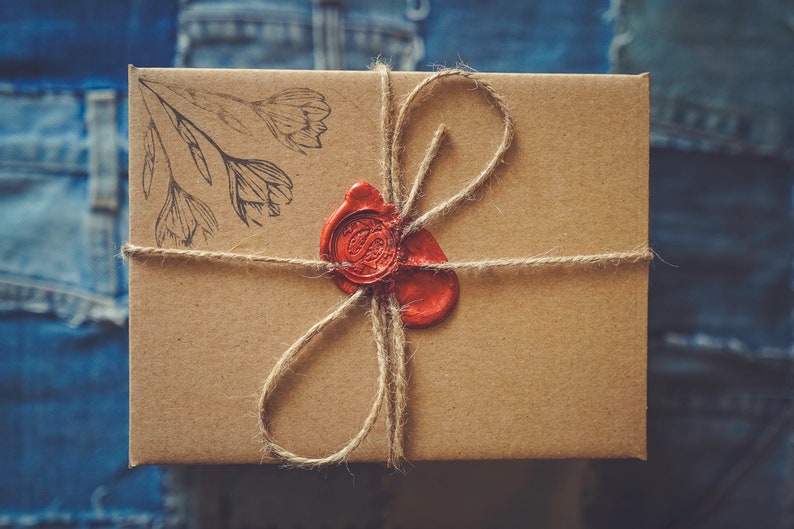 Peut inclure: Une bo&icirc;te cadeau rectangulaire marron attach&eacute;e avec de la ficelle et scell&eacute;e avec un sceau de cire rouge. La bo&icirc;te a un motif floral dans le coin sup&eacute;rieur gauche. Le fond est un tissu en denim bleu.