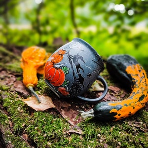 May include: A Halloween-themed ceramic mug with orange pumpkins and a silhouette design. The mug is set on a mossy log with decorative gourds in orange and green. The background is a blurred forest scene.