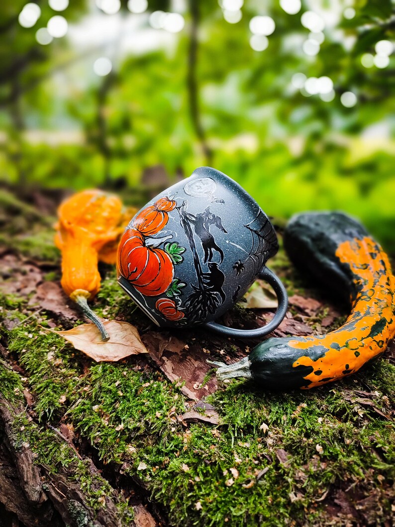 May include: A Halloween-themed ceramic mug with orange pumpkins and a silhouette design. The mug is set on a mossy log with decorative gourds in orange and green. The background is a blurred forest scene.
