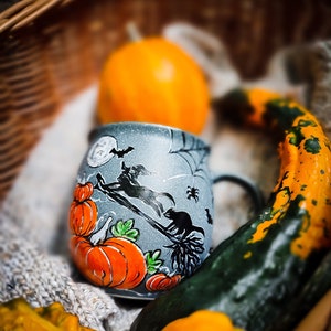 May include: A gray ceramic Halloween mug with orange pumpkins and a witch on a broom design. The mug is inside a wicker basket with a small orange pumpkin and decorative gourds. The mug has a black handle.