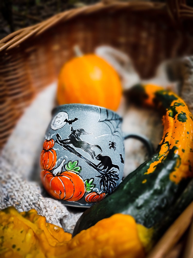 May include: A gray ceramic Halloween mug with orange pumpkins and a witch on a broom design. The mug is inside a wicker basket with a small orange pumpkin and decorative gourds. The mug has a black handle.