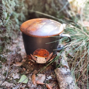 May include: A black ceramic mug with a brown mushroom design and a brown lid. The mug is sitting on a bed of leaves and twigs.