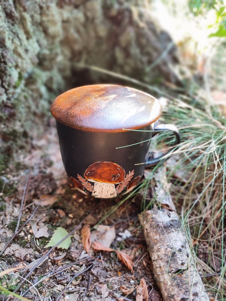May include: A black ceramic mug with a brown mushroom design and a brown lid. The mug is sitting on a bed of leaves and twigs.