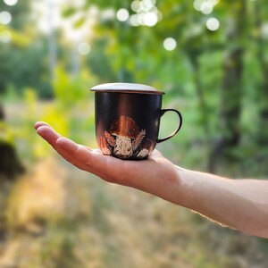 May include: A black ceramic mug with a lid, featuring a mushroom design. The mug is being held in a hand against a blurred background of green foliage.