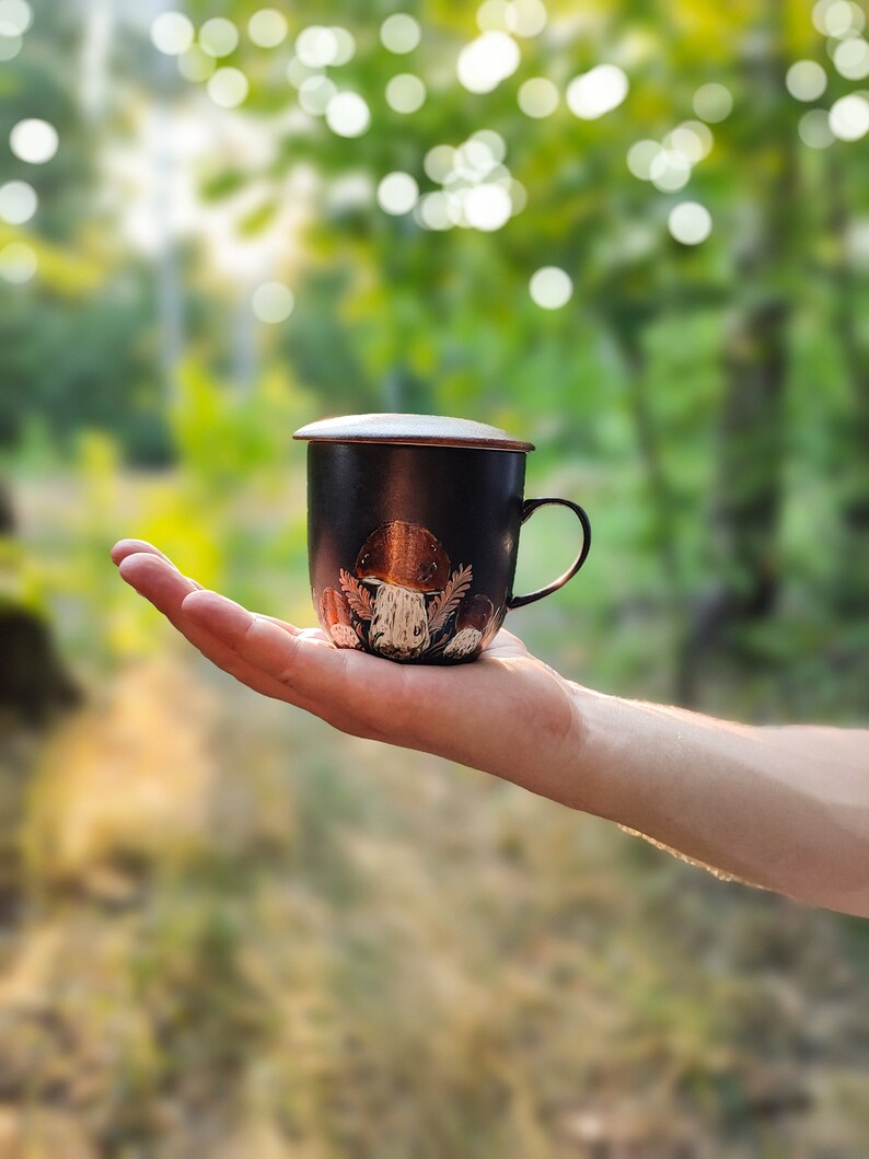 May include: A black ceramic mug with a lid, featuring a mushroom design. The mug is being held in a hand against a blurred background of green foliage.