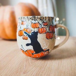 Könnte beinhalten: Eine Keramiktasse mit einer schwarzen Katze, die nach einer Kürbis greift. Die Tasse ist mit einem Muster aus orangefarbenen Kürbissen und Ranken verziert.