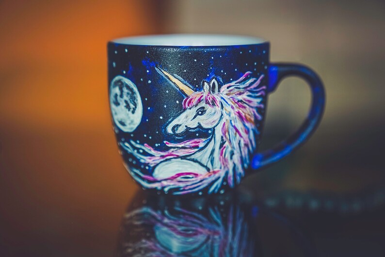 Peut inclure: Tasse en c&eacute;ramique peinte &agrave; la main, avec un fond bleu fonc&eacute;. Elle pr&eacute;sente un motif de licorne avec un corps blanc, une corne dor&eacute;e et une crini&egrave;re rose et violette. Une pleine lune et des &eacute;toiles sont &eacute;galement peintes sur la tasse.