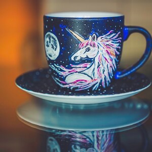 Peut inclure: Un ensemble tasse et soucoupe en c&eacute;ramique peint &agrave; la main. La tasse est bleu fonc&eacute; avec un motif de licorne et de lune blanche. La licorne a une crini&egrave;re rose et violette. La soucoupe est bleu fonc&eacute; avec des points blancs.