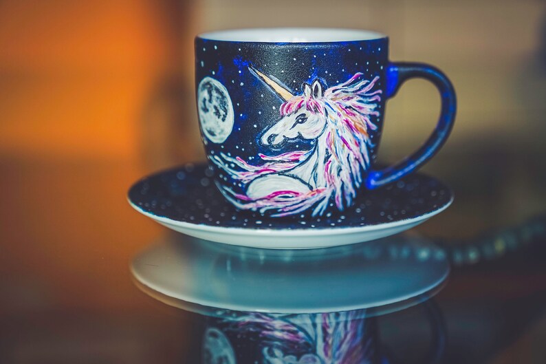 Peut inclure: Un ensemble tasse et soucoupe en c&eacute;ramique peint &agrave; la main. La tasse est bleu fonc&eacute; avec un motif de licorne et de lune blanche. La licorne a une crini&egrave;re rose et violette. La soucoupe est bleu fonc&eacute; avec des points blancs.