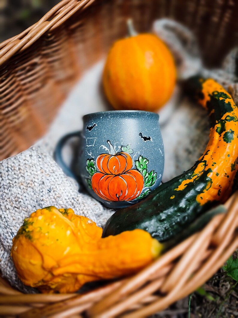 May include: A fall-themed still life featuring a blue mug with a hand-painted pumpkin design, surrounded by gourds and a small pumpkin in a woven basket. The mug has a speckled finish and bat silhouettes. The gourds are orange, yellow, and green.