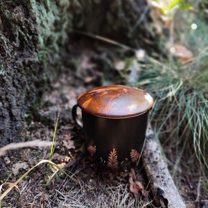 May include: A black ceramic mug with a lid and gold leaf design. The mug is sitting on a bed of brown leaves and grass.