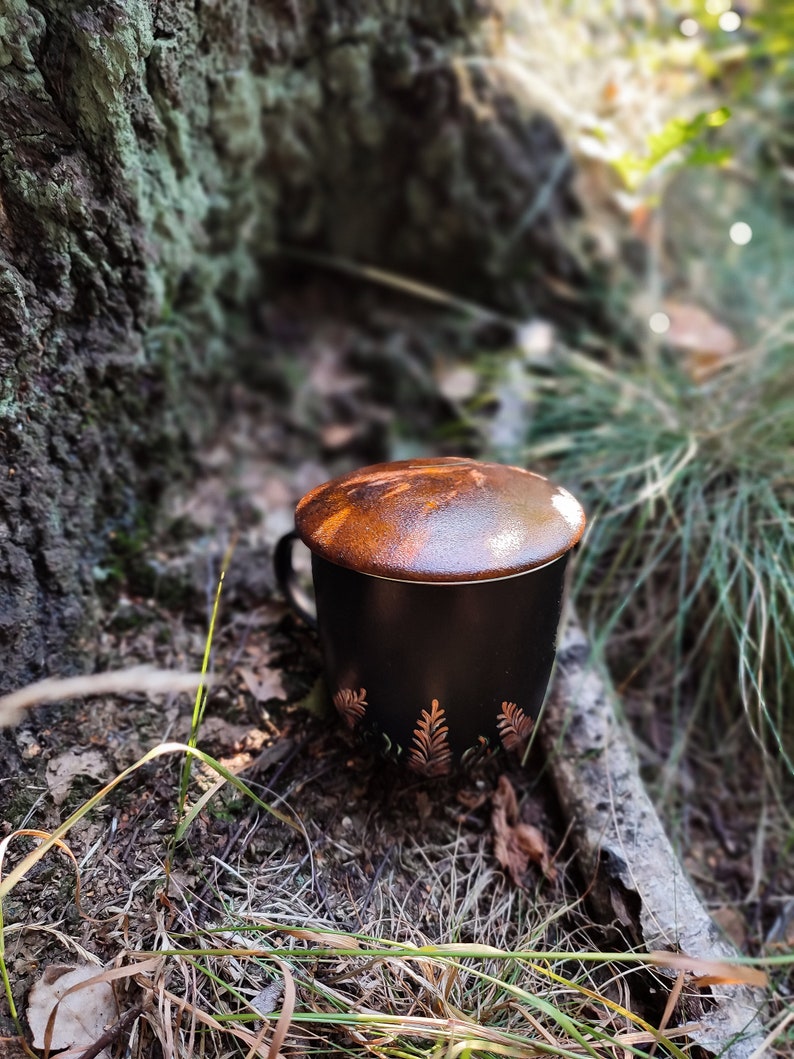 May include: A black ceramic mug with a lid and gold leaf design. The mug is sitting on a bed of brown leaves and grass.