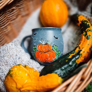 May include: A fall-themed still life featuring a blue mug with a hand-painted pumpkin design, surrounded by gourds and a small pumpkin in a woven basket. The mug has a speckled finish and bat silhouettes. The gourds are orange, yellow, and green.