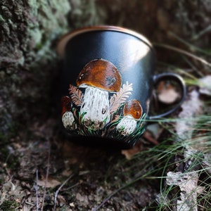 May include: A black ceramic mug featuring a hand-painted design of mushrooms. The mushrooms are brown with white stems and are surrounded by green leaves.
