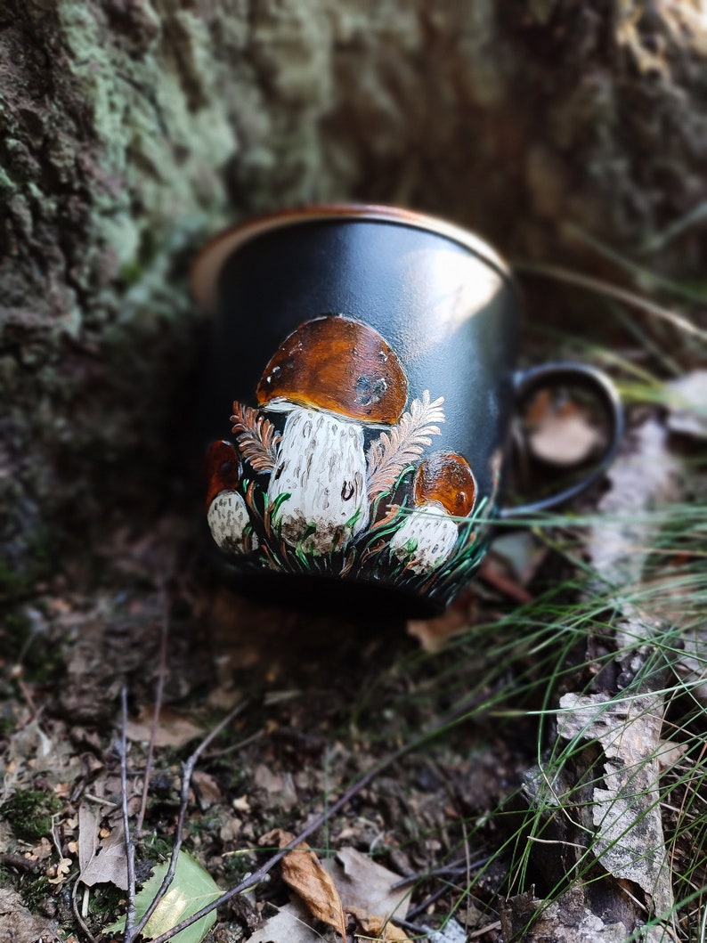 May include: A black ceramic mug featuring a hand-painted design of mushrooms. The mushrooms are brown with white stems and are surrounded by green leaves.
