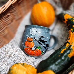 May include: A gray ceramic mug with a Halloween design featuring orange pumpkins, a black witch on a broom, and a full moon. The mug is nestled in a woven basket with gourds and a knitted blanket, creating a fall theme.