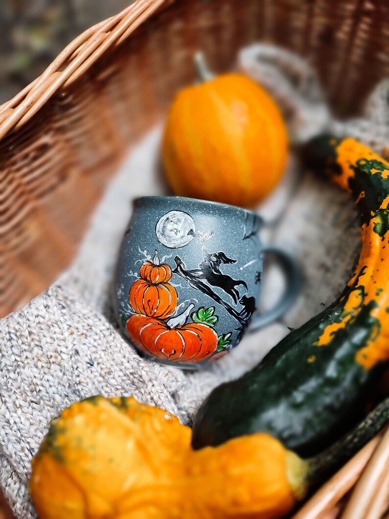 May include: A gray ceramic mug with a Halloween design featuring orange pumpkins, a black witch on a broom, and a full moon. The mug is nestled in a woven basket with gourds and a knitted blanket, creating a fall theme.