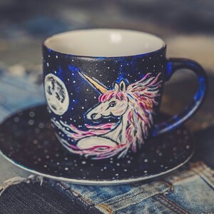 Peut inclure: Un ensemble tasse et soucoupe en c&eacute;ramique peint &agrave; la main. La tasse pr&eacute;sente un motif de licorne avec une corne dor&eacute;e, une crini&egrave;re rose et violette, et une pleine lune sur un fond bleu fonc&eacute; &eacute;toil&eacute;. La soucoupe est bleu fonc&eacute; avec des points blancs.