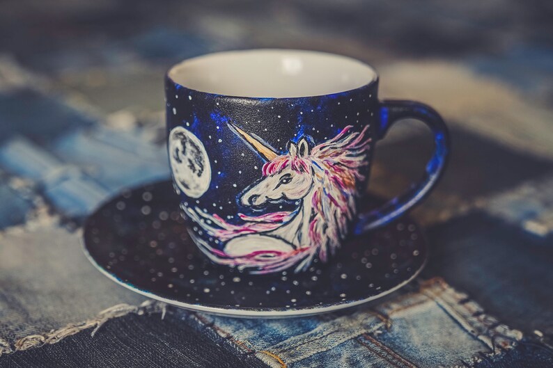 Peut inclure: Un ensemble tasse et soucoupe en c&eacute;ramique peint &agrave; la main. La tasse pr&eacute;sente un motif de licorne avec une corne dor&eacute;e, une crini&egrave;re rose et violette, et une pleine lune sur un fond bleu fonc&eacute; &eacute;toil&eacute;. La soucoupe est bleu fonc&eacute; avec des points blancs.