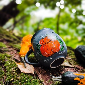 May include: A dark blue ceramic mug with a hand-painted orange pumpkin design. The mug is set on a moss-covered log with small gourds, creating a fall or Halloween theme. The mug has a black handle.