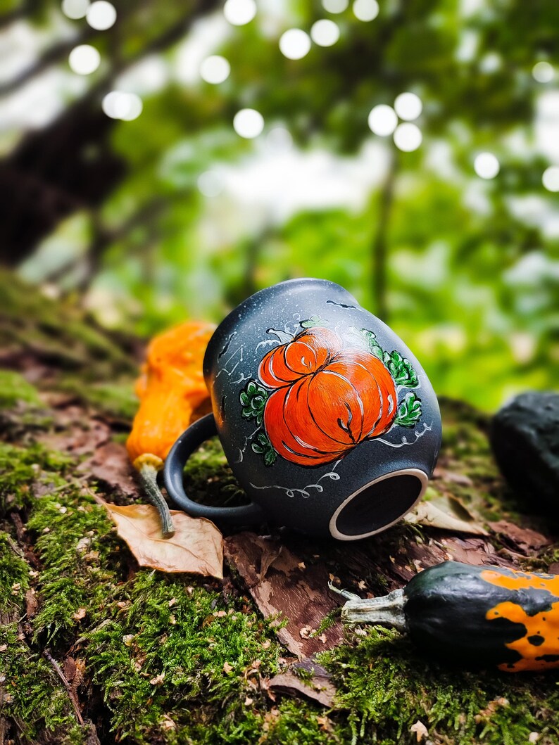 May include: A dark blue ceramic mug with a hand-painted orange pumpkin design. The mug is set on a moss-covered log with small gourds, creating a fall or Halloween theme. The mug has a black handle.