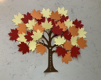 May include: A brown paper tree with a trunk and branches, adorned with colorful maple leaf cutouts in shades of red, orange, and yellow. The word "THANKFUL" is vertically cut out of the tree trunk.