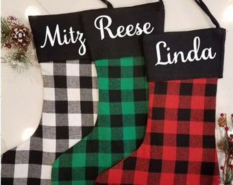 May include: Three Christmas stockings with black tops and plaid patterns in black and white, green and black, and red and black. Each stocking has a name in white script: Mitz, Reese, and Linda. Black hanging loops are attached.