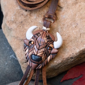 May include: A brown leather cord necklace with a brown and white geometric bison pendant. The pendant is made of polymer clay and has a textured surface.